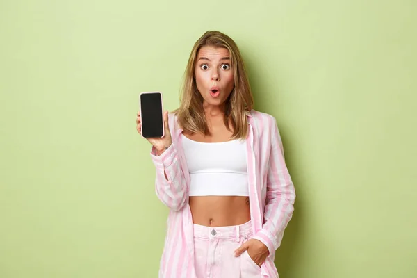 Imagem de modelo feminino atraente loiro em camisa rosa, mostrando tela do smartphone e olhando surpreso, de pé sobre fundo verde — Fotografia de Stock