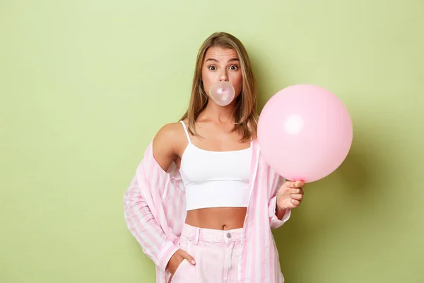 Retrato de menina loira bonita com balão rosa, soprando uma goma de mascar e olhando tolo para a câmera, de pé sobre fundo verde — Fotografia de Stock