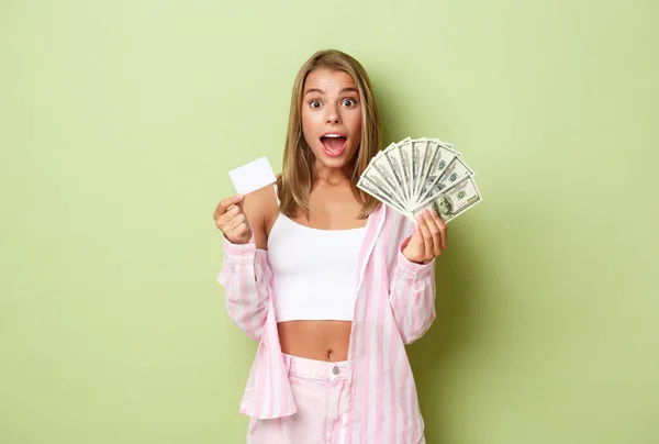 Ritratto di donna bionda eccitata che mostra carta di credito e contanti, in piedi stupito su sfondo verde — Foto Stock