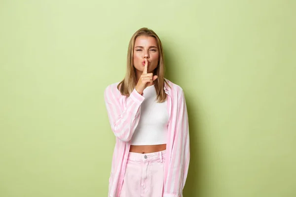 Sensual mujer rubia atractiva en camisa rosa y jeans, escondiendo secreto, haciendo señal de silencio y buscando tortuosa, de pie sobre fondo verde — Foto de Stock