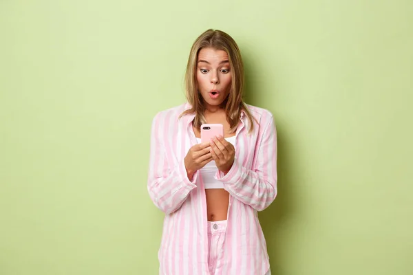 Retrato de menina loira surpreso vestindo camisa rosa, olhando surpreendido para a tela do telefone móvel, reagindo à mensagem ou notificação, de pé sobre fundo verde — Fotografia de Stock