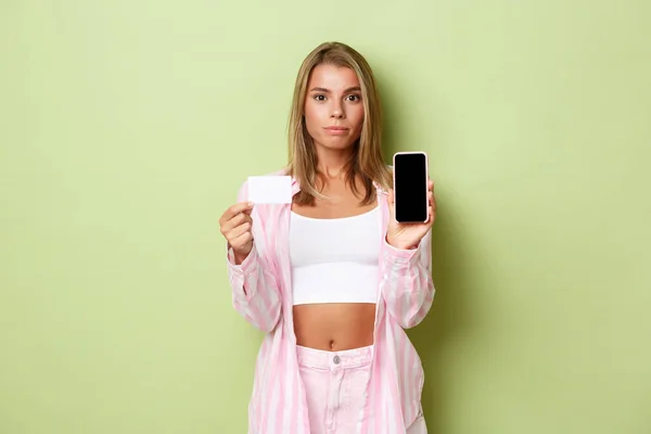 Retrato de mujer rubia confiada en camisa rosa sobre la parte superior de la cosecha, mostrando la pantalla del teléfono inteligente y la tarjeta de crédito, de pie sobre el fondo verde —  Fotos de Stock