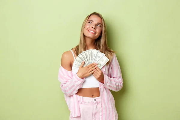 Afbeelding van mooie blonde vrouw in roze shirt, op zoek bedachtzaam op de linker bovenhoek en het houden van geld, dromen over winkelen, staan over groene achtergrond — Stockfoto