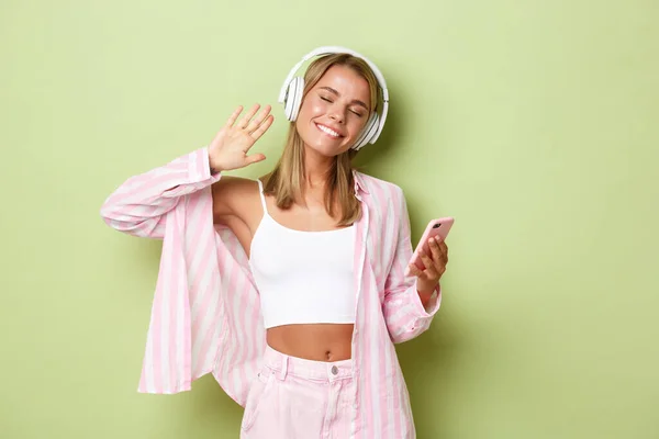 Изображение красивой блондинки в розовой рубашке, слушающей музыку в наушниках и улыбающейся, держащей мобильный телефон, стоящей на зеленом фоне — стоковое фото