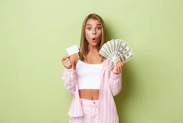 Immagine di donna bionda impressionata ed eccitata, in possesso di contanti e carta di credito, in piedi su sfondo verde — Foto Stock