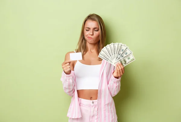 Портрет смущенной блондинки, держащей деньги и нерешительно смотрящей на кредитку, стоящей озадаченной зеленым фоном — стоковое фото