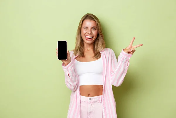 Retrato de menina loira bonito em camisa rosa, mostrando sinal de paz e tela do telefone móvel, sorrindo satisfeito, de pé sobre fundo verde — Fotografia de Stock