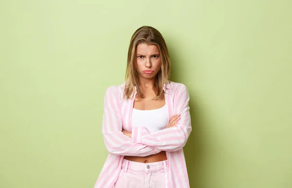 Portrait de fille timide offensée aux cheveux blonds courts, regardant quelqu'un en colère, boudant et croisant les bras sur la poitrine, regardant sous le front en colère, debout sur fond vert — Photo