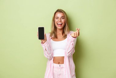 Pembe tişörtlü şık sarışın bir kız resmi, kalp işareti ve cep telefonu ekranı gösteriyor, internette yeşil arka planda duran bir şey gibi.