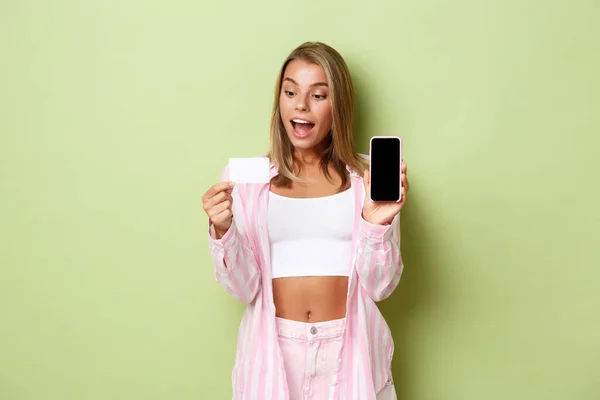 Imagem da menina loira animado em camisa rosa, mostrando cartão de crédito e tela do telefone móvel, de pé sobre fundo verde — Fotografia de Stock