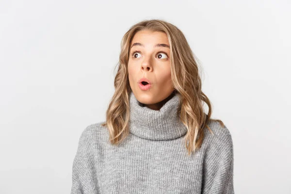 Close-up of pretty blond girl in grey sweater, looking at upper left corner with surprised face, gasping fascinated, standing over white background — Stock Photo, Image