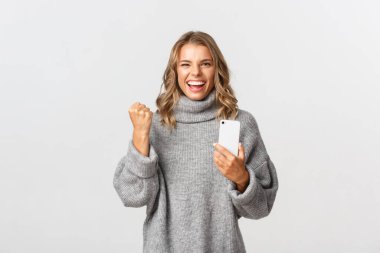 Gri kazaklı mutlu bir kız resmi, evet diyor ve zafer kazanıyor, cep telefonunu tutuyor, akıllı telefon uygulamasında hedefe ulaşıyor, beyaz arka planda duruyor.