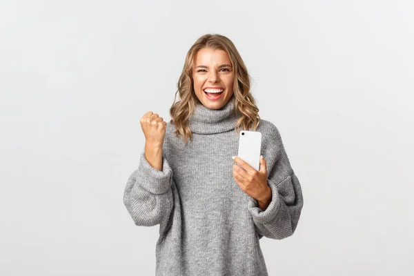 Bild av lycklig vinnande flicka i grå tröja, säger ja och triumferande, håller mobiltelefon, uppnå mål i smartphone ansökan, står över vit bakgrund — Stockfoto