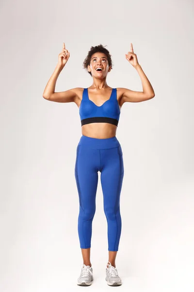 Comprimento total de mulher afro-americana animado e feliz em sportswear para treino, olhando e apontando os dedos com o rosto surpreso, fundo branco — Fotografia de Stock