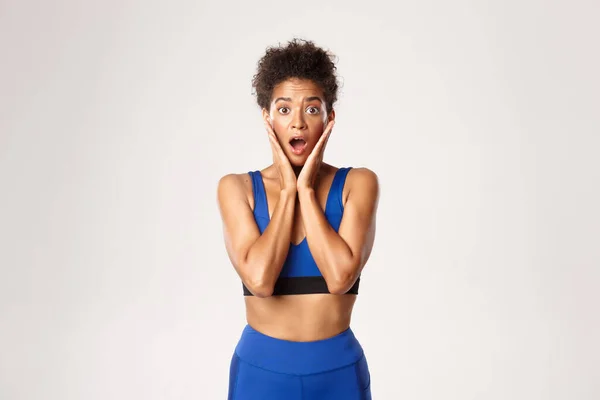 Waist-up van verrast Afrikaans-Amerikaanse fit vrouwelijke atleet, op zoek verbaasd, open mond verwonderd, staande tegen een witte achtergrond in blauwe sport outfit — Stockfoto