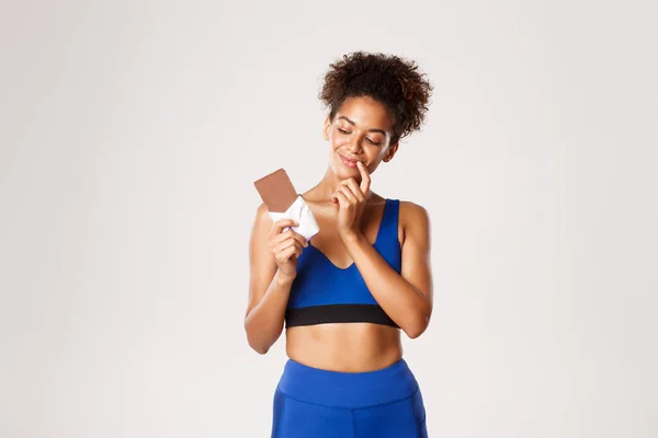 Tevreden Afrikaans-Amerikaanse fitness vrouw in blauwe sport kostuum, op zoek naar chocolade bar met verleid gezicht, staande tegen een witte achtergrond — Stockfoto