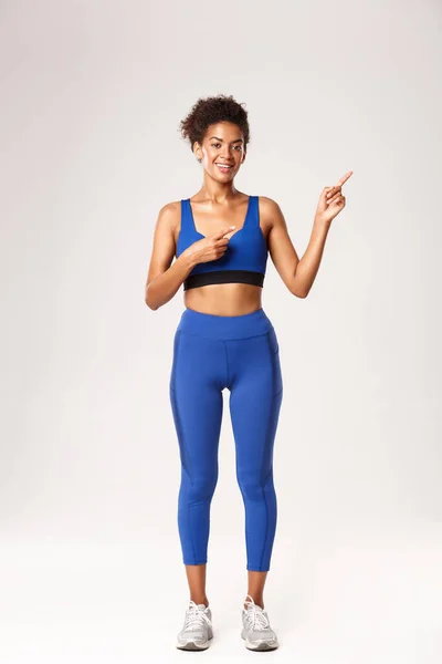 Comprimento total de atraente sorridente mulher afro-americana com cabelo penteado, vestindo sportswear azul, apontando os dedos no canto superior direito, mostrando promo fitness, fundo branco — Fotografia de Stock