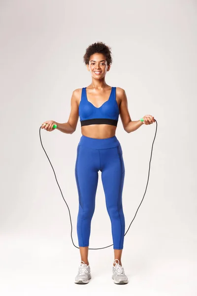 Longitud completa de atleta femenina fuerte en traje deportivo azul, saltando con saltar la cuerda y sonriendo, haciendo ejercicios sobre fondo blanco — Foto de Stock