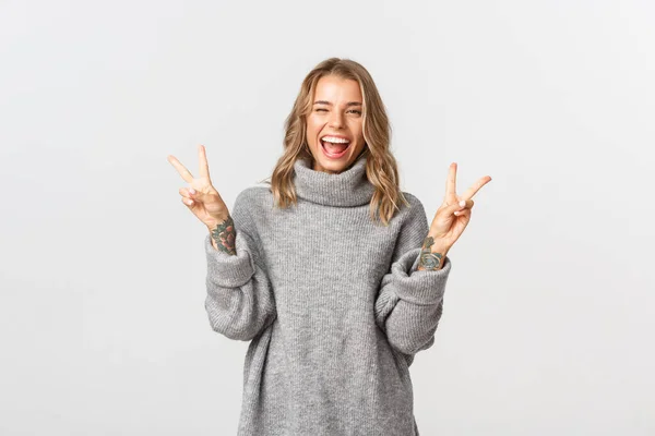 Primer plano de la atractiva chica rubia en suéter gris, mostrando signos de paz y sonriendo, de pie sobre fondo blanco —  Fotos de Stock