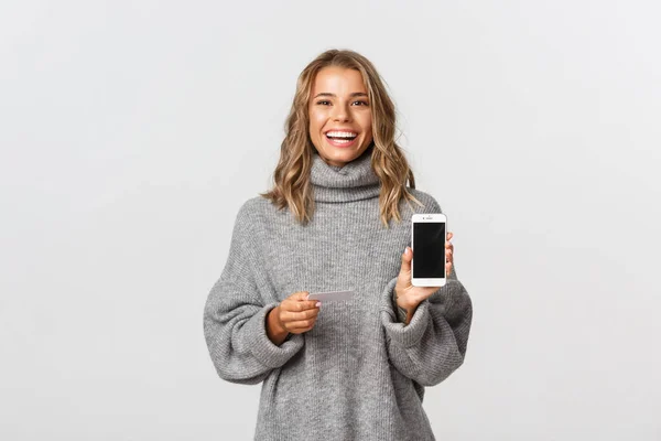 Atractiva rubia en suéter gris que muestra la pantalla del teléfono móvil y la tarjeta de crédito, sonriendo a la cámara, de pie sobre fondo blanco — Foto de Stock