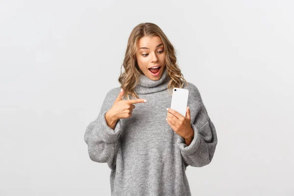 Retrato de modelo femenino caucásico atractivo en suéter gris, mirando sorprendido y apuntando al teléfono móvil, recomiendan algo, fondo blanco — Foto de Stock