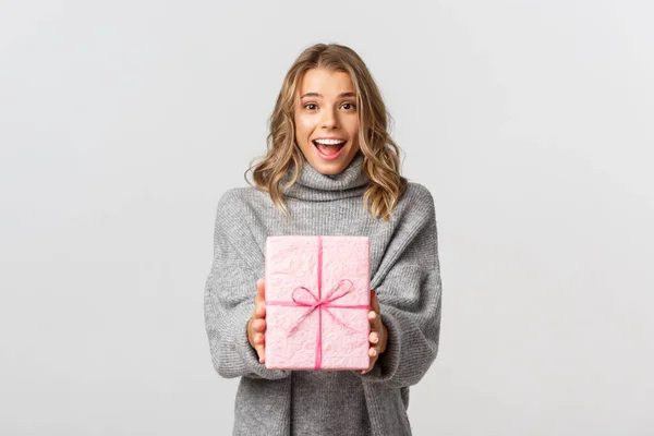 Hermosa mujer joven en suéter gris, dando regalo, felicitando a alguien con cumpleaños, de pie sobre fondo blanco —  Fotos de Stock