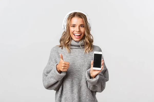 Imagem da menina loira satisfeita olhando feliz, mostrando polegares para cima, ouvir música em fones de ouvido e demonstrar tela do smartphone, fundo branco — Fotografia de Stock