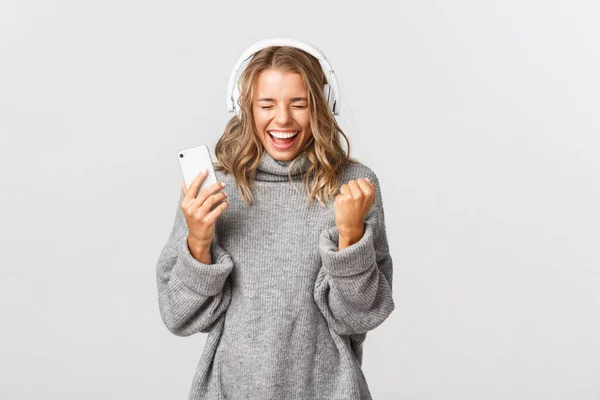 Söt vinnande flicka i grå tröja, lyssna musik i hörlurar, hålla smartphone och skriker av glädje, står över vit bakgrund — Stockfoto