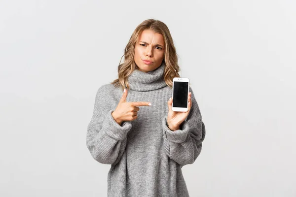 Retrato de menina irritada apontando o dedo para o smartphone, mostrando algo decepcionante e esperando explicação, de pé sobre fundo branco — Fotografia de Stock