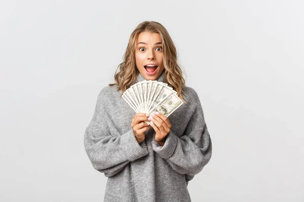 Afbeelding van opgewonden aantrekkelijke meisje in grijze trui, met geld en kijken verbaasd, staande over witte achtergrond — Stockfoto