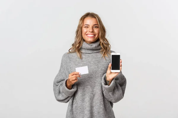 Hermosa modelo femenina rubia en suéter gris, que muestra la tarjeta de crédito y la pantalla del teléfono móvil, de pie sobre fondo blanco — Foto de Stock