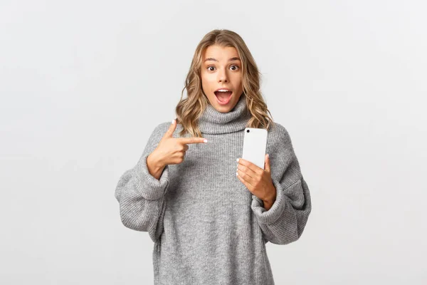 Porträtt av förbluffad blond flicka i grå tröja, peka finger åt mobiltelefon, rekommenderar titta på något online, står över vit bakgrund — Stockfoto