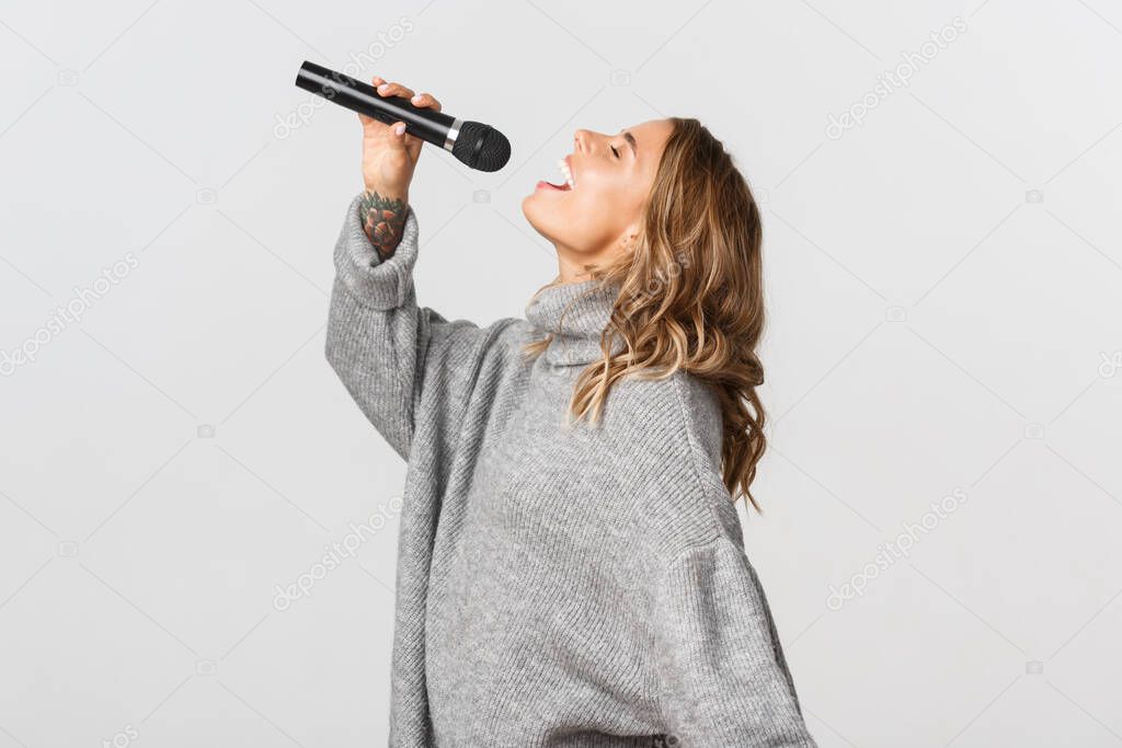 Profile shot of happy blond girl singing in microphone. Girl performing a song in karaoke, standing over white background