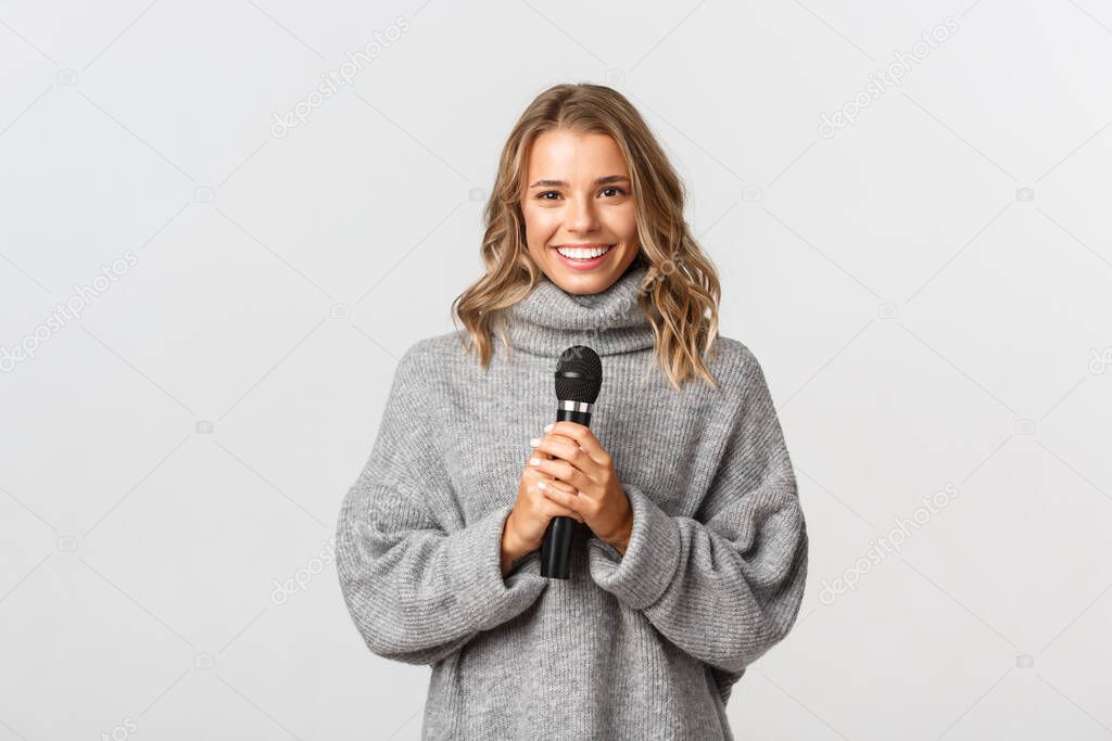 Portrait of beautiful blond girl in grey sweater, smiling while standing with microphone against white background