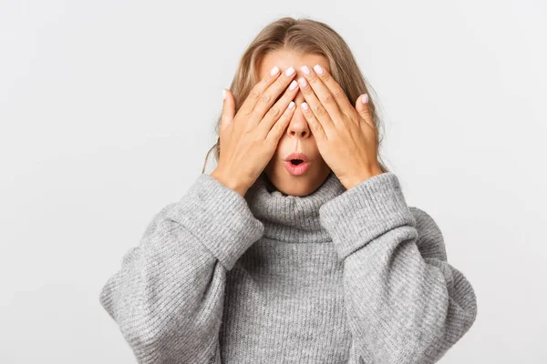 Close-up of happy beautiful woman in grey sweater, standing blindsided with hands on face, waiting for surprise, white background — Stock Photo, Image