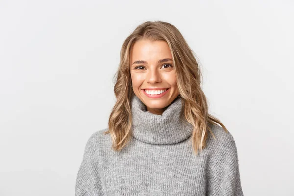 Primer plano de la rubia feliz en suéter gris, mirando a la cámara y sonriendo, de pie sobre fondo blanco —  Fotos de Stock