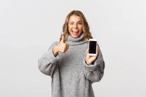 Bild av attraktiv blond flicka i grå tröja, ser nöjd och visar något bra på smartphone skärm, göra tummen upp i godkännande, vit bakgrund — Stockfoto