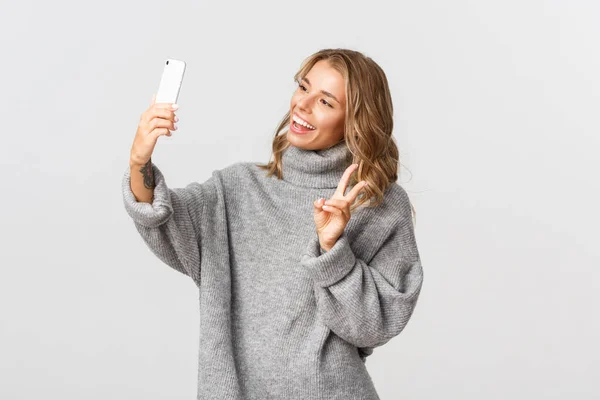 Porträtt av attraktiv ung blond flicka i grå tröja håller smartphone, visar fred tecken och ta selfie, står över vit bakgrund — Stockfoto
