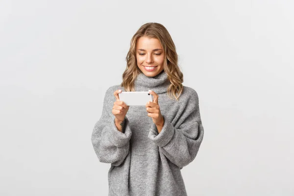 Bild av attraktiv blond flicka som håller mobiltelefon horisontellt som om titta på något eller spela spel, stående över vit bakgrund — Stockfoto
