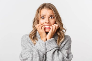 Gri kazaklı, tırnak yiyen, dehşete düşmüş, beyaz arka planda duran korkmuş sarışın kadının yakın çekimi.