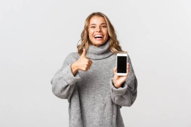 Gri kazaklı, mutlu bir kadının resmi, gülüyor ve gülümsüyor, başparmağını kaldırıyor ve cep telefonu ekranını gösteriyor, beyaz arka planda duruyor.