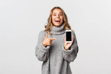 Gri kazaklı mutlu beyaz kız portresi, komik bir şeye gülüyor, akıllı telefon ekranını işaret ediyor, beyaz arka planda duruyor.