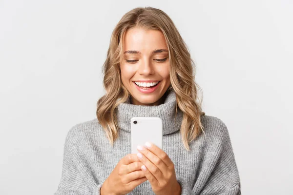 Närbild av attraktiv blond flicka i grå tröja, kontrollera meddelanden på telefon eller handla online, titta på smartphone med glad ansikte, står över vit bakgrund — Stockfoto