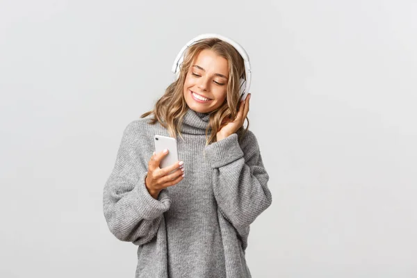 Hermosa chica rubia en suéter gris, escuchando música en auriculares y sosteniendo el teléfono móvil, de pie sobre fondo blanco — Foto de Stock