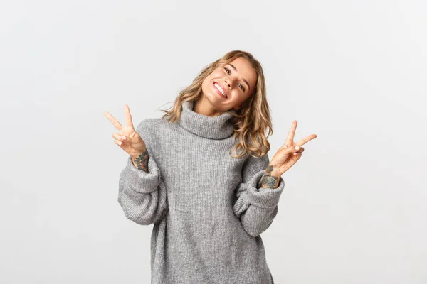 Close-up of attractive blond girl in grey sweater, showing peace signs and smiling, standing over white background — Stock Photo, Image