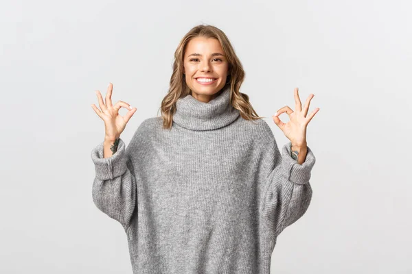 Jovem mulher satisfeita com cabelo curto loiro, vestindo camisola cinza, mostrando sinais ok e sorrindo satisfeito, de pé sobre fundo branco — Fotografia de Stock