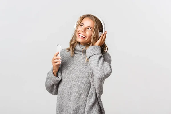 Ung attraktiv blond flicka lyssnar musik i trådlösa hörlurar, ler och tittar på övre vänstra hörnet, håller mobiltelefon — Stockfoto