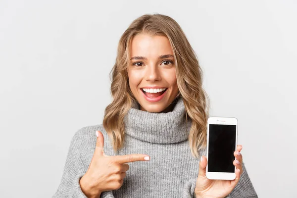 Närbild av attraktiv blond flicka i grå tröja, pekar finger på mobiltelefon skärm, står över vit bakgrund — Stockfoto