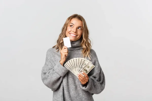 Bella ragazza bionda dall'aspetto premuroso e sorridente, in possesso di carta di credito e denaro, in piedi su sfondo bianco — Foto Stock