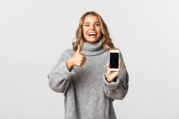 Bild av nöjd lycklig kvinna i grå tröja, skrattar och ler, visar tummen upp och mobiltelefon skärm, står över vit bakgrund — Stockfoto